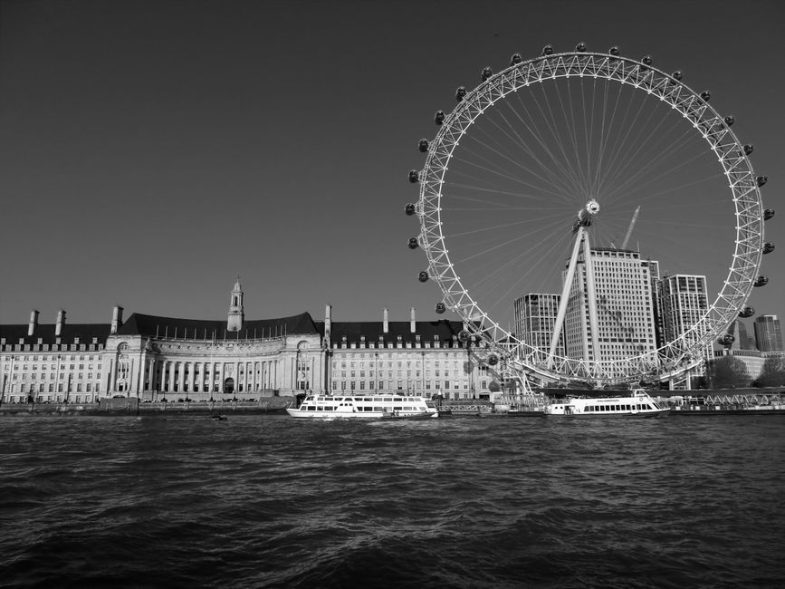 A black and white photograph capturing the London Eye Ferris wheel situated on the South Bank of the River Thames, with several passenger boats docked along the water's edge. In the background, there