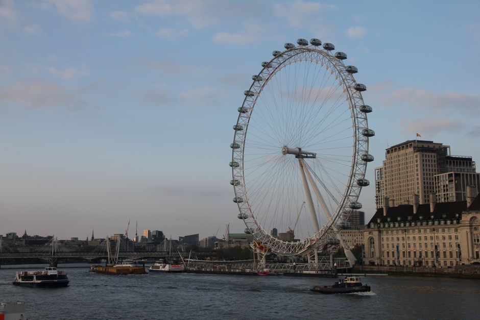 A black and white photograph capturing the London Eye Ferris wheel situated on the South Bank of the River Thames, with several passenger boats docked along the water's edge. In the background, there